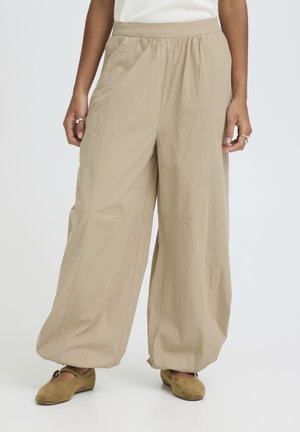 Žena v béžových širokých nohaviciach s elastickým pásom a béžovými semišovými topánkami Mary Jane, stojaca pred jednoduchým pozadím.