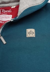 Teal ribbad tröja med grå krage, som har en silverdragkedja och en fyrkantig beige etikett med svart text och korslagda redskap.