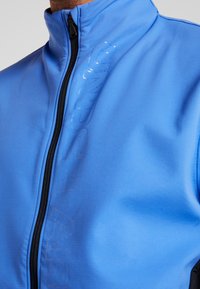 Blauwe zip-up jack met een hoge kraag, gestructureerde stof en een zwarte rits; heeft een subtiel reliëflogo aan de voorkant.