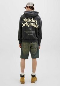 Dark gray hoodie with "Studio Originals" printed in large cream letters on the back, paired with green-toned denim shorts and brown boots.