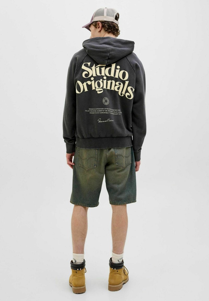 Dark gray hoodie with "Studio Originals" printed in large cream letters on the back, paired with green-toned denim shorts and brown boots.
