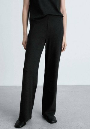 Czarne spodnie o szerokich nogawkach zestawione z czarnymi butami o kwadratowym nosku, noszone przez osobę stojącą na szarej podłodze na tle jasnoszarym.