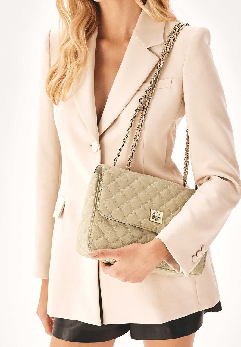 Kevyesti tikattu vaaleanvihreä käsilaukku, jossa ketjuhihna, kullanvärinen metallikoristelu ja läppäkiinnitys. Yhdistetty beigen värisen bleiserin kanssa.