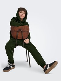 Hoodie vert foncé et pantalon de survêtement assorti, assis sur une chaise en bois. Baskets noires avec des accents blancs et une semelle texturée.