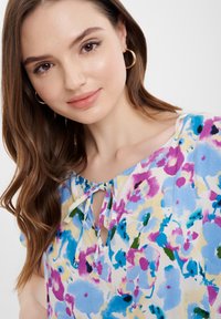 Blumenbluse mit Krawattenkragen, aus leichtem Material gefertigt. Hat ein farbenfrohes Muster aus blauen, lila und grünen Blumen auf einem hellen Hintergrund.
