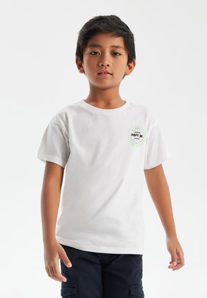 T-shirt en coton blanc à manches courtes, col rond, avec un logo vert sur la poitrine. porté avec un short cargo sombre.