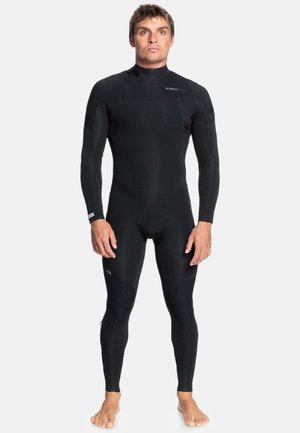 Hombre de pie descalzo vistiendo un traje de neopreno negro completo, mirando hacia adelante con los brazos relajados a los lados contra un fondo liso.