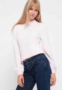 Gaiši rozā adīta džemperis ar augstu apkakli un pūkainām garām piedurknēm, apvienots ar tumši ziliem džinsiem. Ir ribotas aproces un apakšmalas.