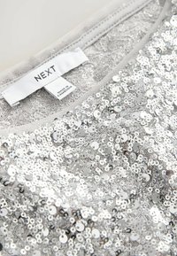Top di paillettes argento con una texture stampata, dotato di un'etichetta con "NEXT" e "Made in Cambodia". Finitura lucida con dettagli riflettenti.