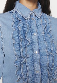 Ljusblå denimskjorta med volangdetaljer längs knäppningen, försedd med silverfärgade tryckknappar och en klassisk krage. Strukturerad yta.