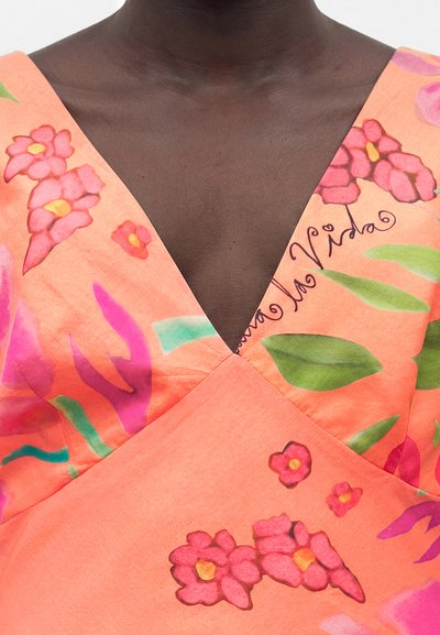 Φόρεμα κοραλί με βαθύ V-λαιμόκοψη, ροζ και κόκκινα λουλουδάτα σχέδια, πράσινα φύλλα και κείμενο με καλλιγραφικά γράμματα στην περιοχή του στήθους.