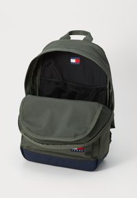 Green fabric backpack with a fold-over flap, navy bottom, and black mesh pocket. Features a logo patch on the front and padded handle.