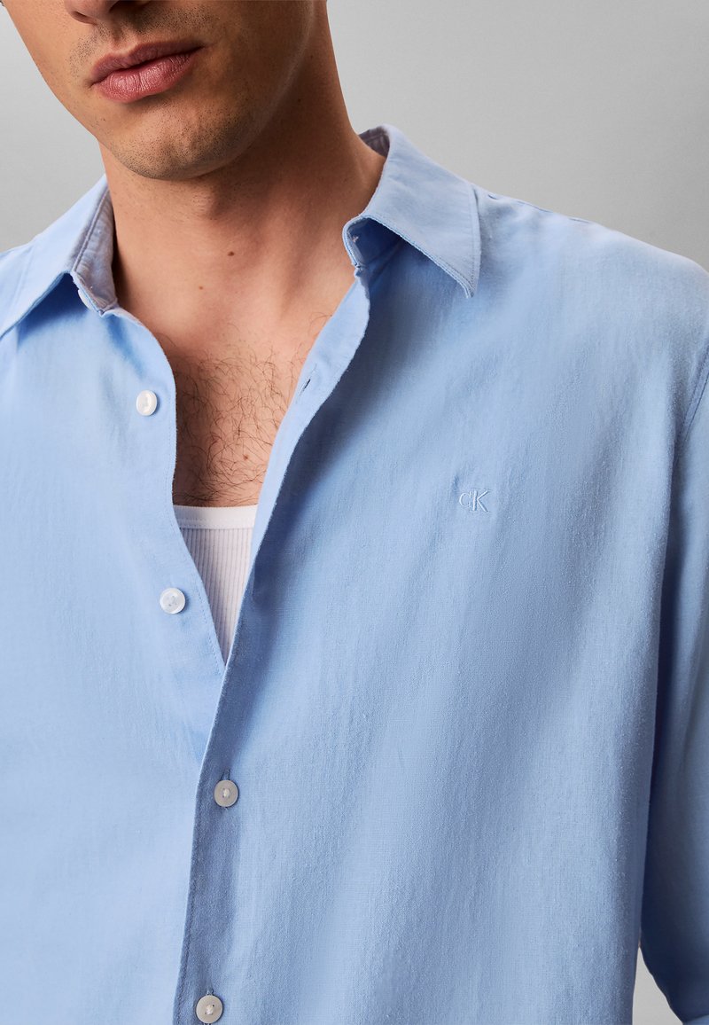 Man draagt lichtblauw overhemd met knopen over een witte geribbelde tanktop, toont wat borsthaar en gedeeltelijk gezicht met gesloten lippen.