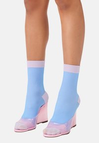 Transparente rosa High-heel Sandalen mit offenen Zehen, kombiniert mit ribbeligen hellblauen Socken mit einem lilafarbenen Bündchen. Glatte Textur, modernes Design.