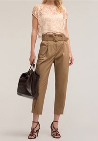 Femme portant un top court en dentelle beige, un pantalon taille haute tan ceinturé, des talons strappy marron foncé, et tenant un sac à main en cuir marron foncé.
