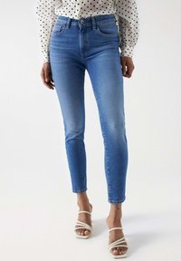 Jean en denim bleu taille haute avec une coupe ajustée, présentant un design délavé et un léger effet de frottement, associé à des sandales beiges à lanières.