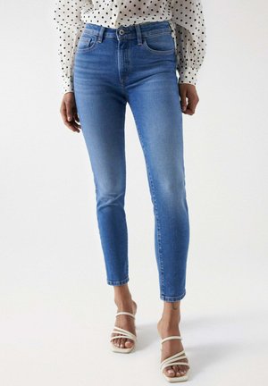 Jean en denim bleu taille haute avec une coupe ajustée, présentant un design délavé et un léger effet de frottement, associé à des sandales beiges à lanières.