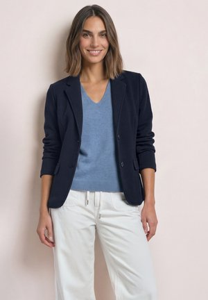 Donna con capelli castani fino alle spalle che indossa un blazer nero, maglione blu con scollo a V e pantaloni bianchi, sorridente su uno sfondo uniforme.