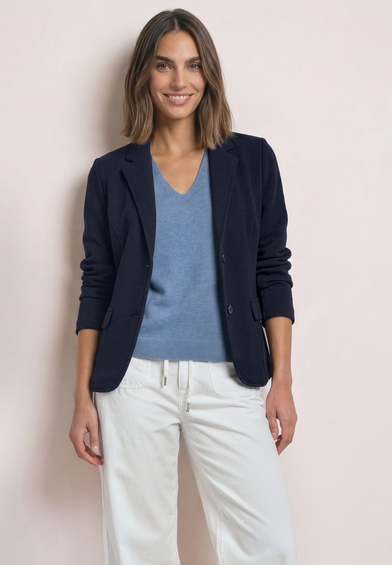 Femme aux cheveux bruns mi-longs portant un blazer noir, un pull bleu col en V et un pantalon blanc, souriante devant un fond uni.