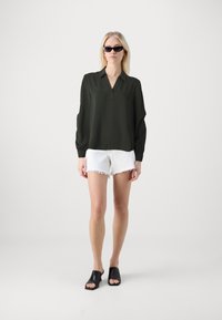 Blusa verde scuro con colletto e maniche ampie, abbinata a pantaloni corti bianchi sfilacciati e mule nere con punta aperta. Tessuto di texture liscia.