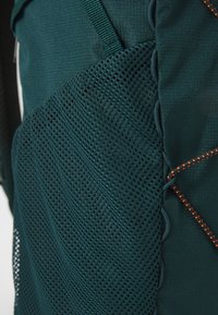 Teksturirana tamnozelena tkanina s mrežastim panelom i narančastim šavovima. Karakteristike uključuju vertikalne linije i mrežu prozračnih otvora.