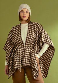Écharpe en damier marron et beige avec une ouverture à l'avant, ceinturée à la taille. Portée par-dessus un pull à côtes léger, accessoirisé d'un bonnet en tricot léger.