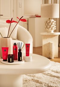 Czerwone produkty kosmetyczne i czarna świeca umieszczone na białej marmurowej tacy na okrągłym stole, obok w beżowym wazonie znajdują się czerwone kwiaty.
