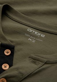 Oliivinvihreä puuvillapaidan T-paita, jossa on pyöreä pääntie, musta nappilista ja etiketti, jossa on kankaan sisältö ja kokotiedot.