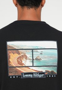 Sort t-shirt med et grafisk print af en naturskøn kystvej og havudsigter i tre paneler, med "Tommy Hilfiger" tekst og etableringsdato.