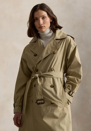 Mladá žena s hnedými vlasmi po ramená, oblečená v béžovom trenčkotu cez biely rolák, pózujúca s jednou rukou v vrecku na neutrálnej pozadí.