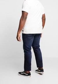 Dark blue jeans with a slim fit, showing back pockets and a subtle fade. Paired with black sneakers featuring white accents.