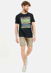 T-shirt en coton marine avec un graphique 'Vers de Nouveaux Horizons' en vert, beige et bleu ; associé à un short beige et des baskets claires.