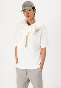Weißes Baumwoll-T-Shirt kombiniert mit hellgrauen, strukturierten Hosen. Ein cremefarbener Pullover ist über die Schultern gelegt. Das Model trägt eine beige Mütze.