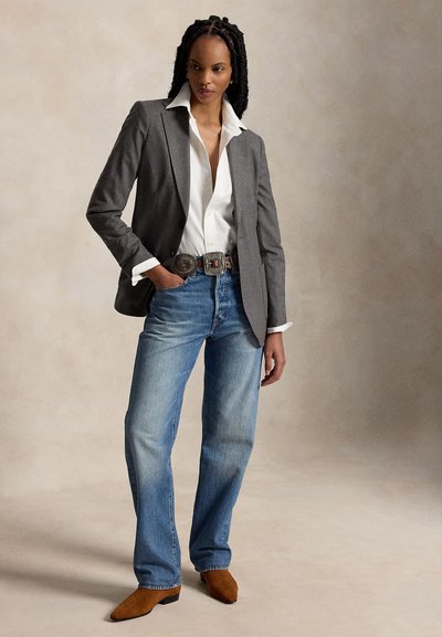 Žena v šedém saku, bílém tričku, modrých džínách, ozdobném pásku a hnědých špičatých botách stojí s jednou rukou v kapse.