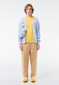 Lichtblauwe cardigan over een gele T-shirt, beige broeken en gele sneakers. Inclusief een lichtblauwe schoudertas met een gladde textuur.