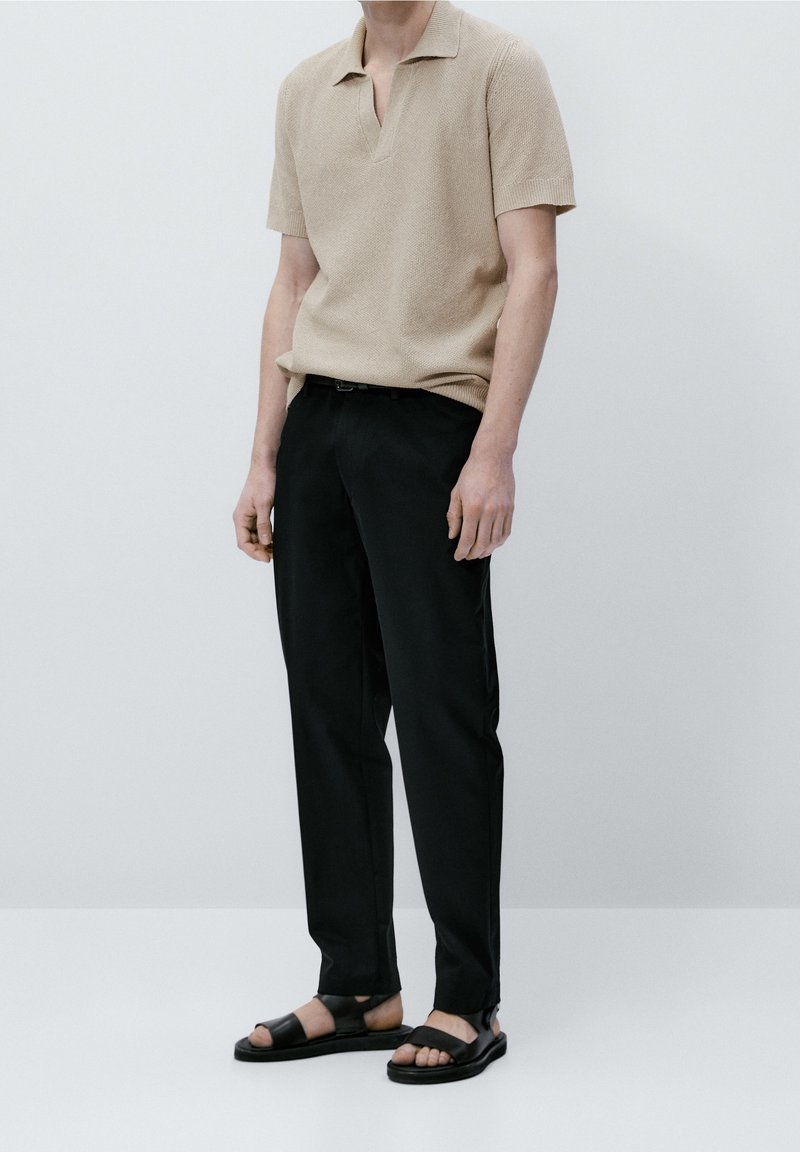 Beige neulepikeepaita, jossa on lyhyt kaulus, yhdistettynä mustiin housuihin ja mustiin sandaaleihin, mattapintainen ja suora leikkaus.