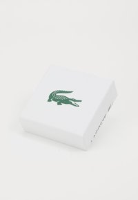 Boîte rectangulaire blanche avec une surface texturée. Logo de crocodile vert sur le dessus, avec "LACOSTE" imprimé en vert sur le côté.