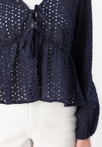 Námornícky modrá blúzka z očkovanej látky s peplum lemom, šnúrkou na krku a balónovými rukávmi. Nosené na bielych nohaviciach.