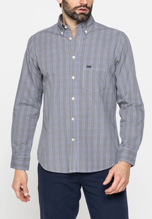 Uomo che indossa una camicia a quadri grigia e blu con maniche lunghe e pantaloni scuri, in piedi davanti a uno sfondo bianco.