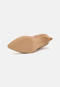 Beige ruskind pumper med spids tå, en glat og struktureret overdel samt en slank, tapet hæl. Gummisålen har vandrette riller for bedre greb.