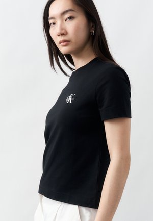Ung kvinde iført sort Calvin Klein T-shirt og hvide bukser, stående mod en ensartet lys baggrund og kigger let til siden.