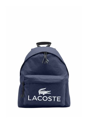 Mochila azul marino con bolsillo frontal con cremallera y asa negra, con el logo blanco de Lacoste con un cocodrilo encima del nombre de la marca.