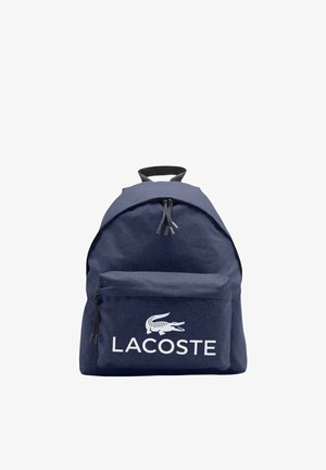 Mochila azul marino con bolsillo frontal con cremallera y asa negra, con el logo blanco de Lacoste con un cocodrilo encima del nombre de la marca.