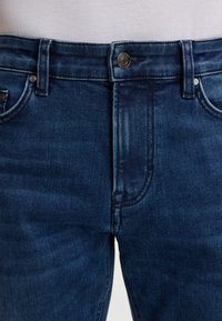 Jeans en denim bleu avec une taille mi-haute, dotés d'une fermeture à bouton en métal, d'un design à cinq poches et d'un léger délavage sur le tissu.