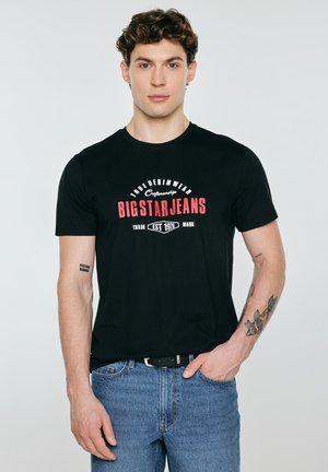 Młody mężczyzna z kręconymi włosami, ubrany w czarną koszulkę "Big Star Jeans" oraz niebieskie dżinsy, stoi z jedną ręką w kieszeni na jednolitym tle.