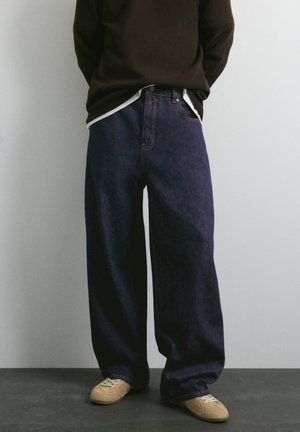 Personne portant un jean large bleu foncé, un pull foncé et des baskets beiges, debout avec les mains dans le dos.