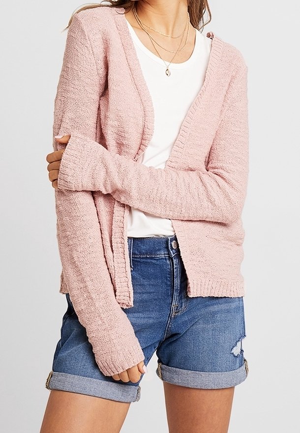 Růžový pletený cardigan s texturovaným povrchem, otevřený vpředu, dlouhými rukávy a žebrovanými manžetami, doplněný bílým tričkem a džínovými šortkami.