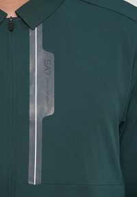 Πράσινο αθλητικό σακάκι με φερμουάρ, διαθέτει ανακλαστική ετικέτα με λογότυπο και υφή, τονισμένο με δομημένο γιακά.