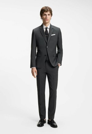 Ung mand iført et tætsiddende sort jakkesæt med hvid skjorte, sort slips, lommetørklæde og sorte pæne sko foran en ensfarvet hvid baggrund.