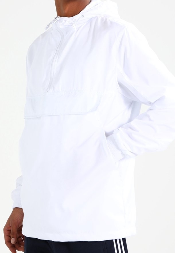 Personne portant une veste coupe-vent blanche à capuche avec fermeture éclair à l'avant et les mains dans les poches, associée à un pantalon foncé.